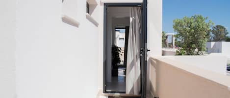 Une porte ouverte donnant sur un balcon, d’où l’on peut admirer un arbre et un bâtiment en arrière-plan.