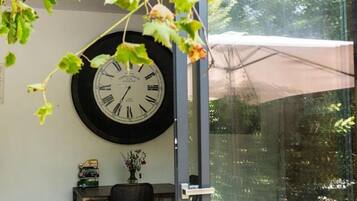 Um quarto com um relógio de parede grande, uma mesa pequena com um vaso de flores e uma janela com vista para o verde.