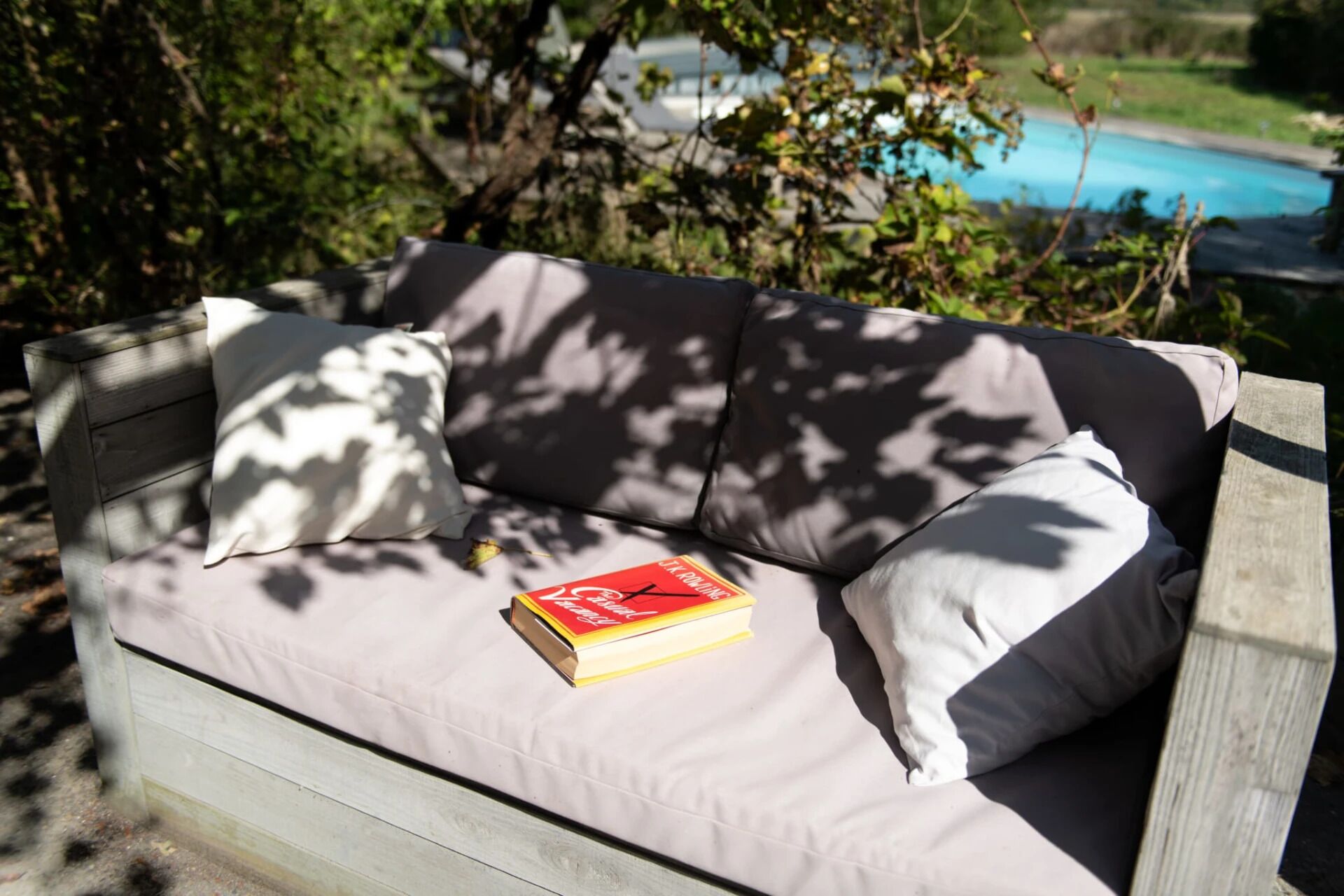 A wooden outdoor sofa with cushions, a book, and a pool in the background.