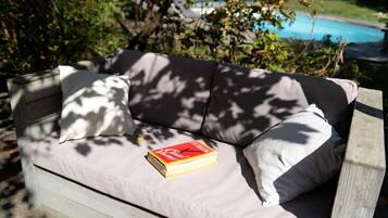 A wooden outdoor sofa with cushions, a book, and a pool in the background.