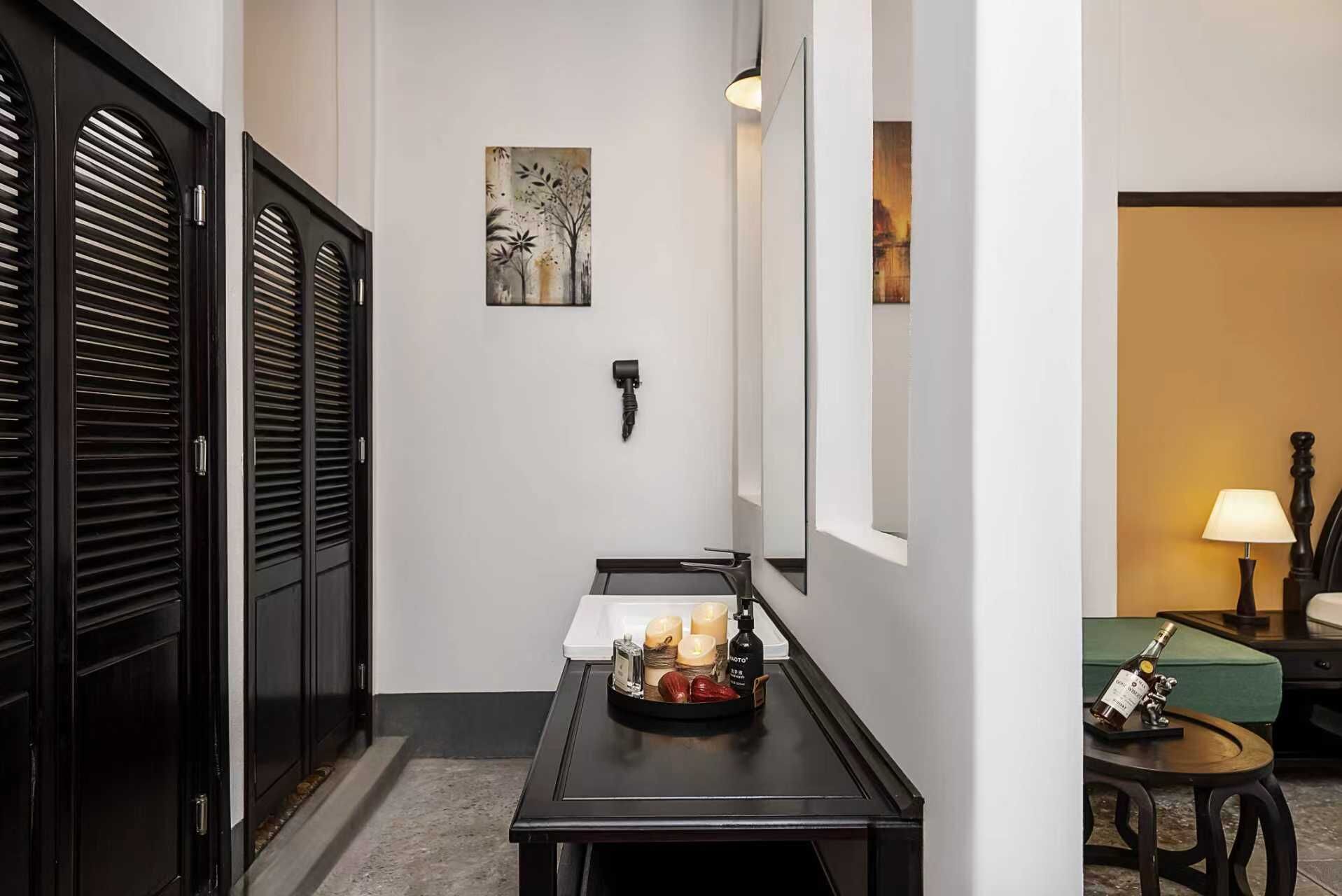 A hallway with a black door, a wall-mounted light fixture, a small table with candles and a bottle, and a painting on the wall.