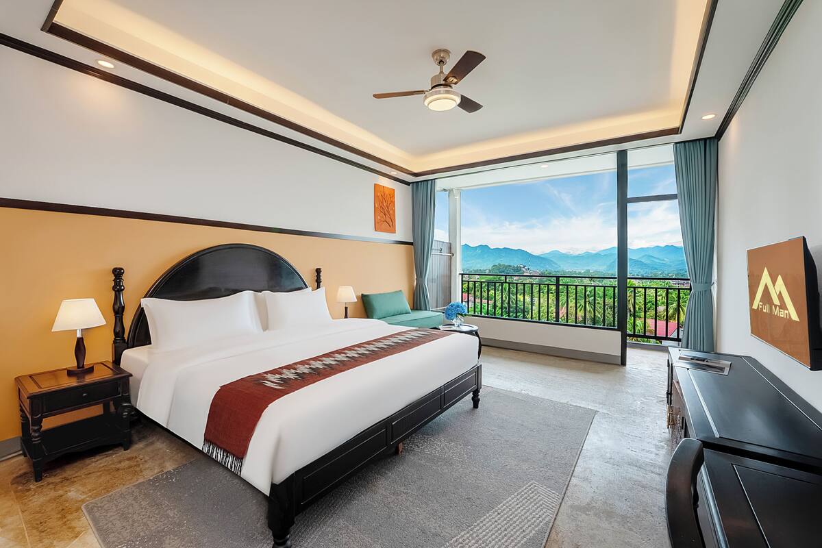 A hotel room with a large bed, a ceiling fan, a view of mountains, and a balcony with a railing.