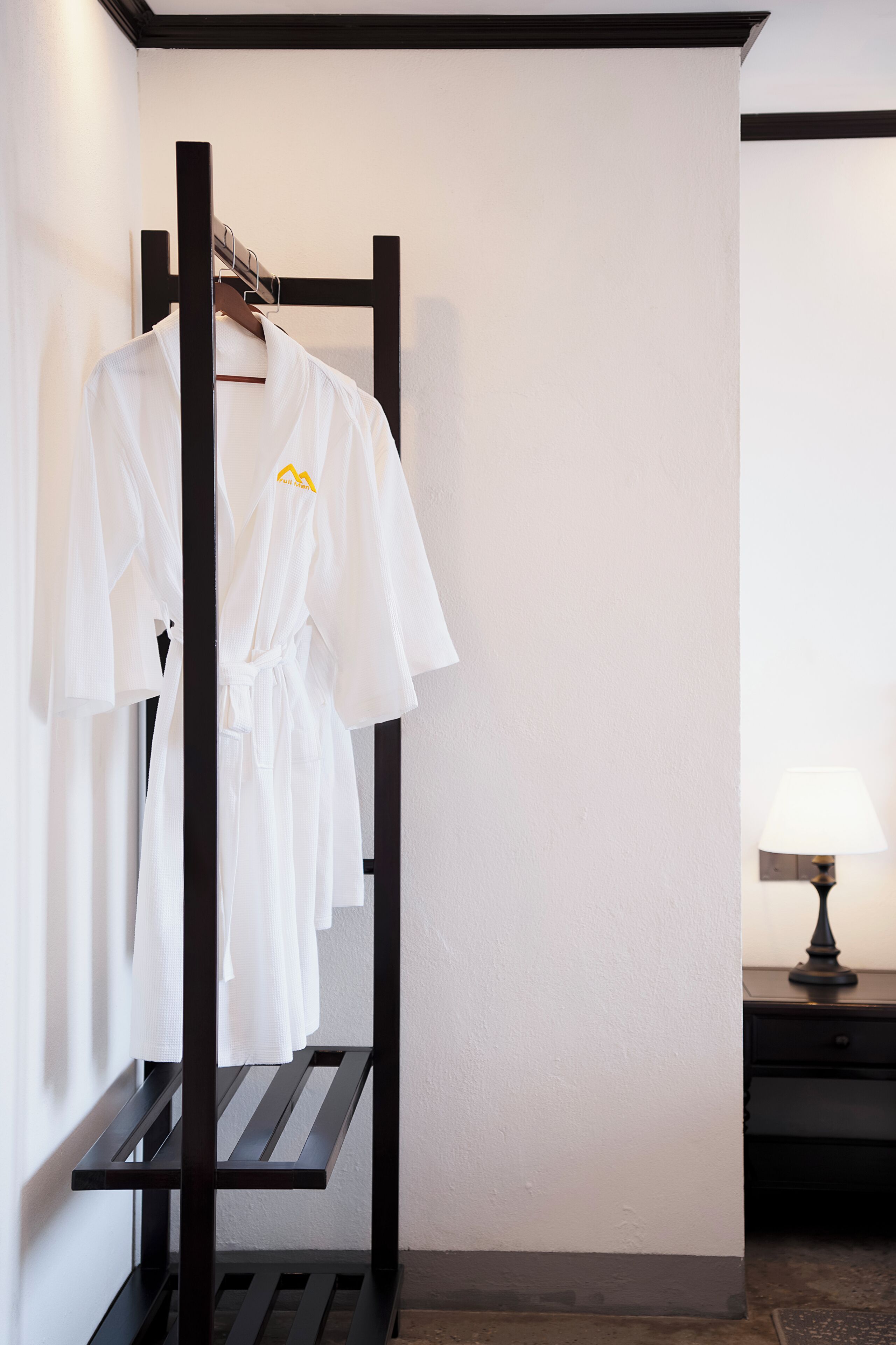 A room with a black metal rack holding white robes, a lamp on a bedside table, and a wooden bench.