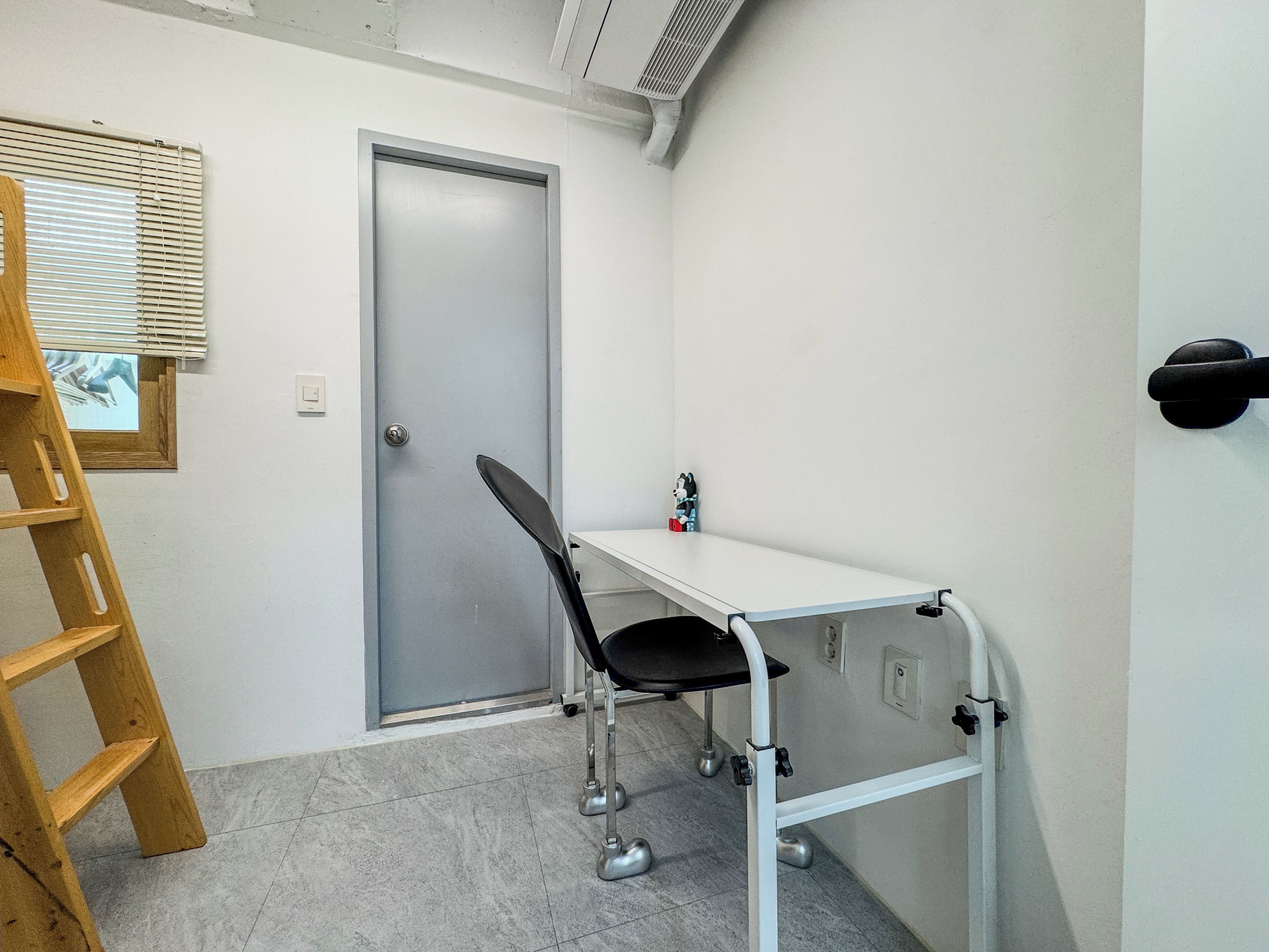 A room with a white desk, a chair, a wooden ladder, a door, and a window with blinds.