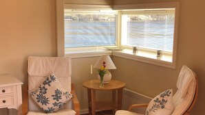 A room with two wooden chairs with white cushions and floral patterns, a small wooden table with a lamp and vase, and a view of the water through the window. - Ramsvig Handelsstad (Stavanger)