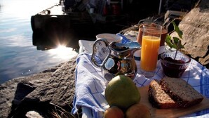 A picnic setup with a metal pitcher, glass of orange juice, bowl of jam, and bread on a blanket by a body of water. - Ramsvig Handelsstad (Stavanger)