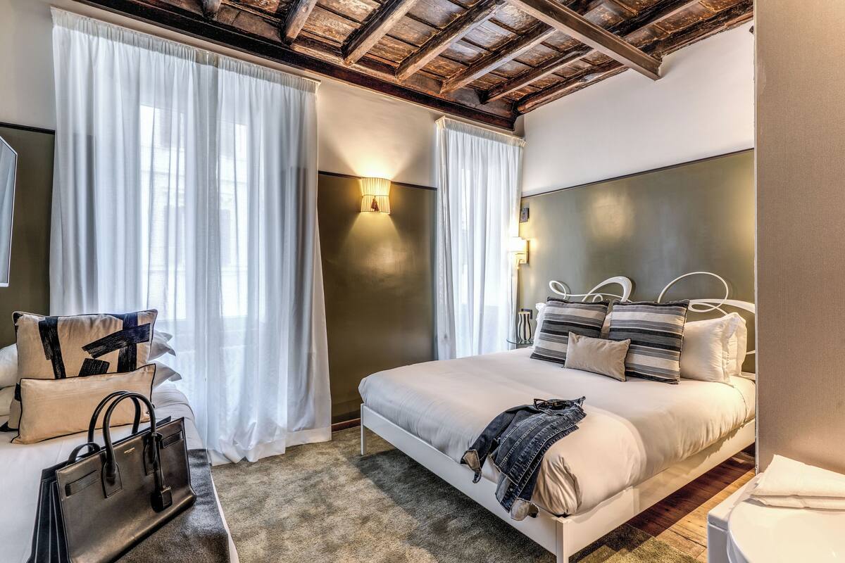 A hotel room with a large bed, white curtains, a wooden ceiling, and a nightstand with a handbag.