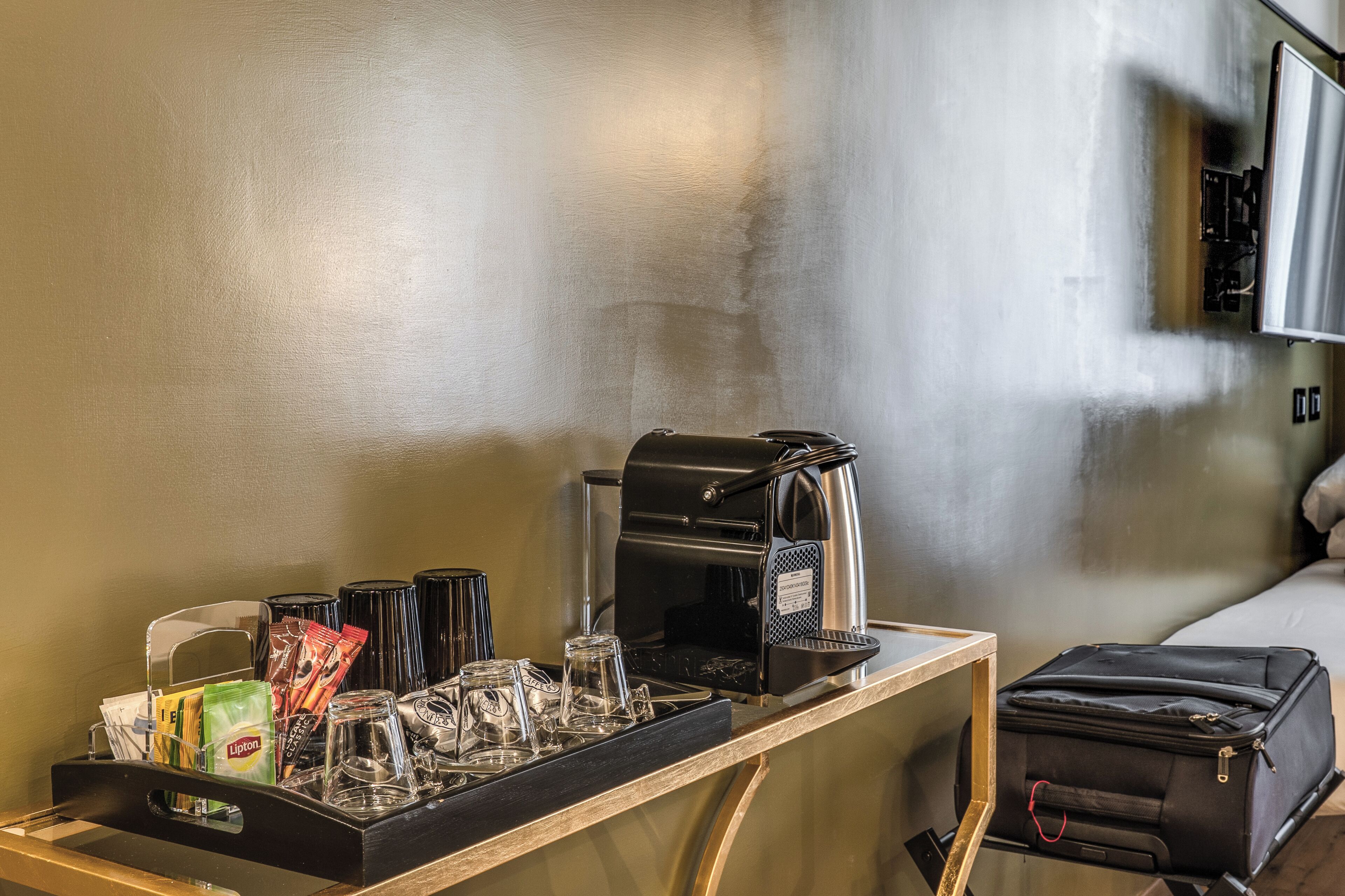 A hotel room with a coffee station, a suitcase, and a bed.