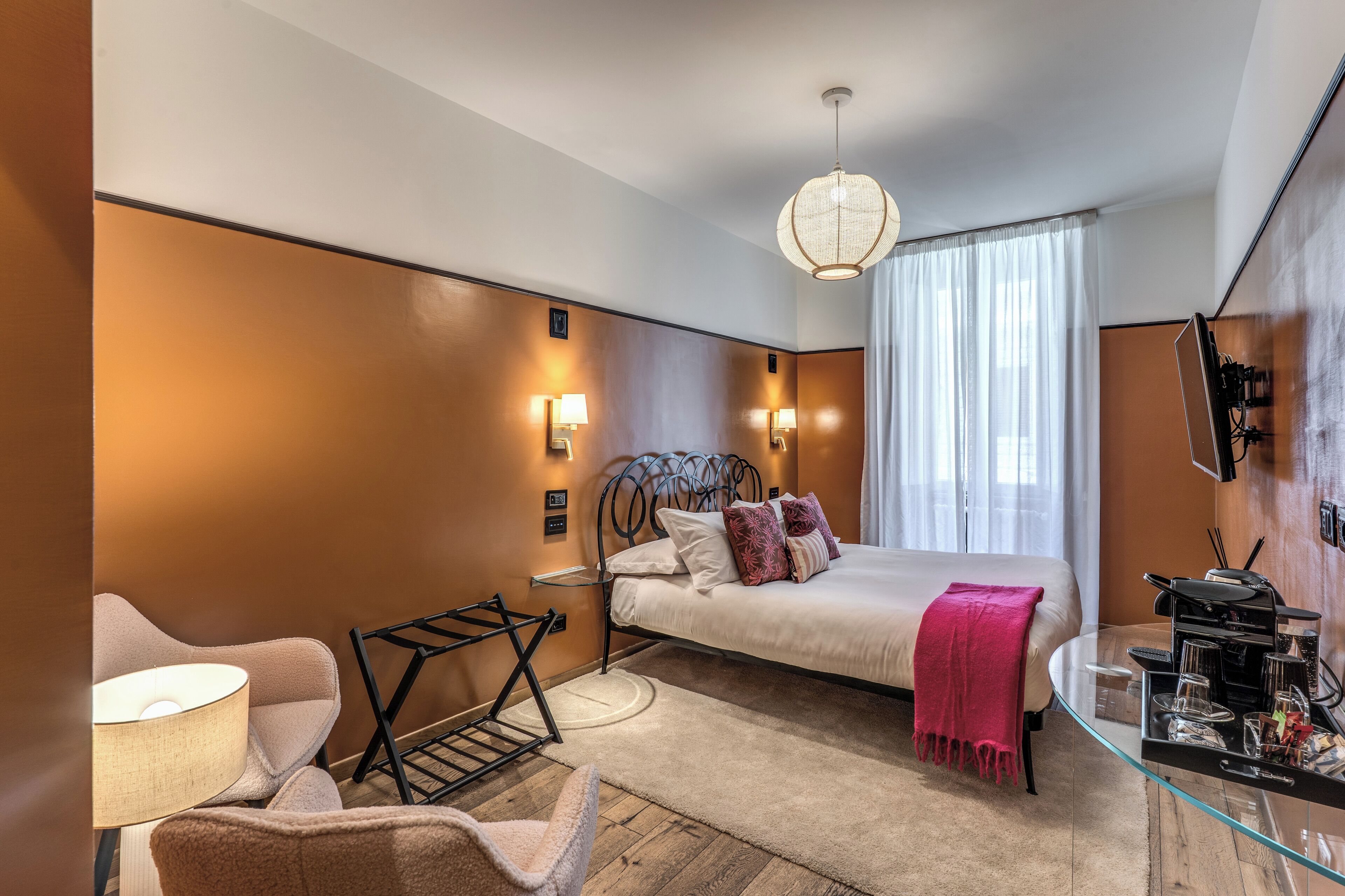 A modern hotel room with a bed, a glass side table, a chair, a lamp, and a hanging light fixture.