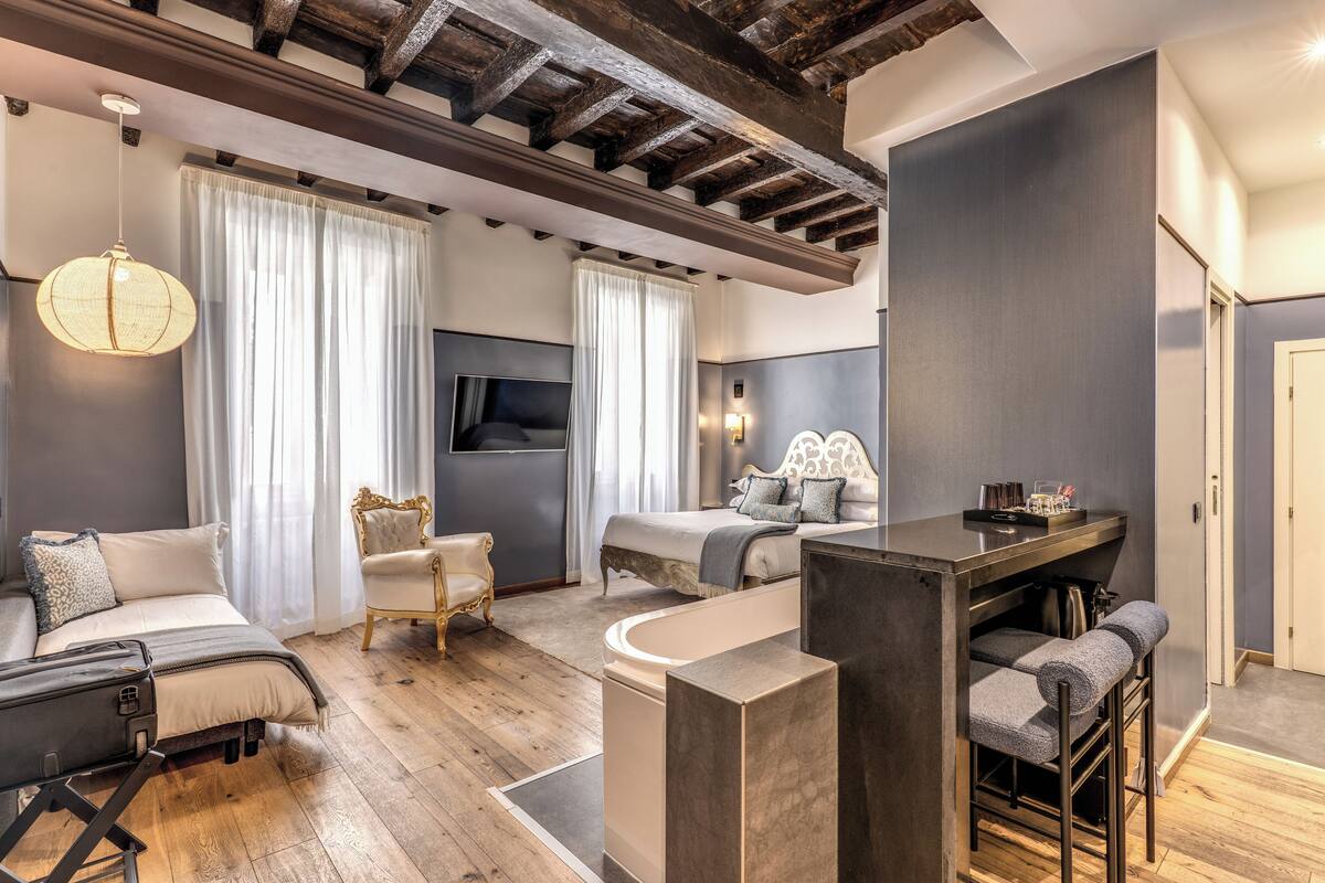 A modern hotel room with a wooden ceiling, a large bed, a sitting area with a sofa and armchair, a small bar area, and a television.