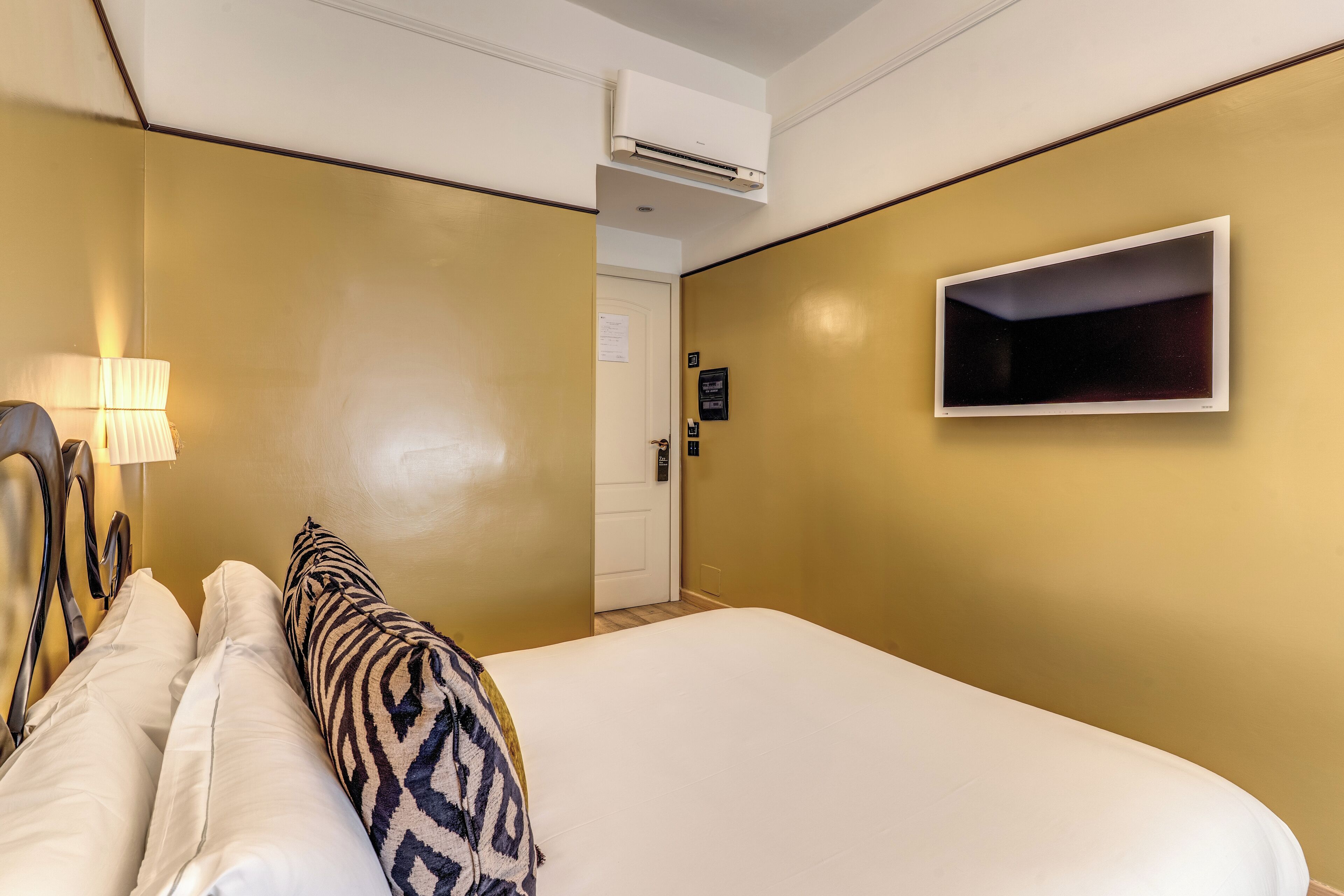 A hotel room with a bed, a TV mounted on the wall, a lamp, and an air conditioning unit.