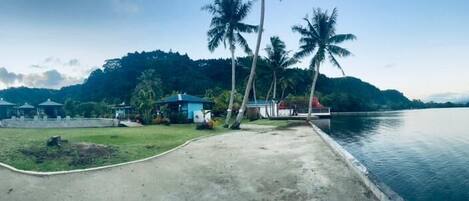 A coastal resort with a concrete walkway leading to a dock, surrounded by palm trees and a mountainous backdrop.