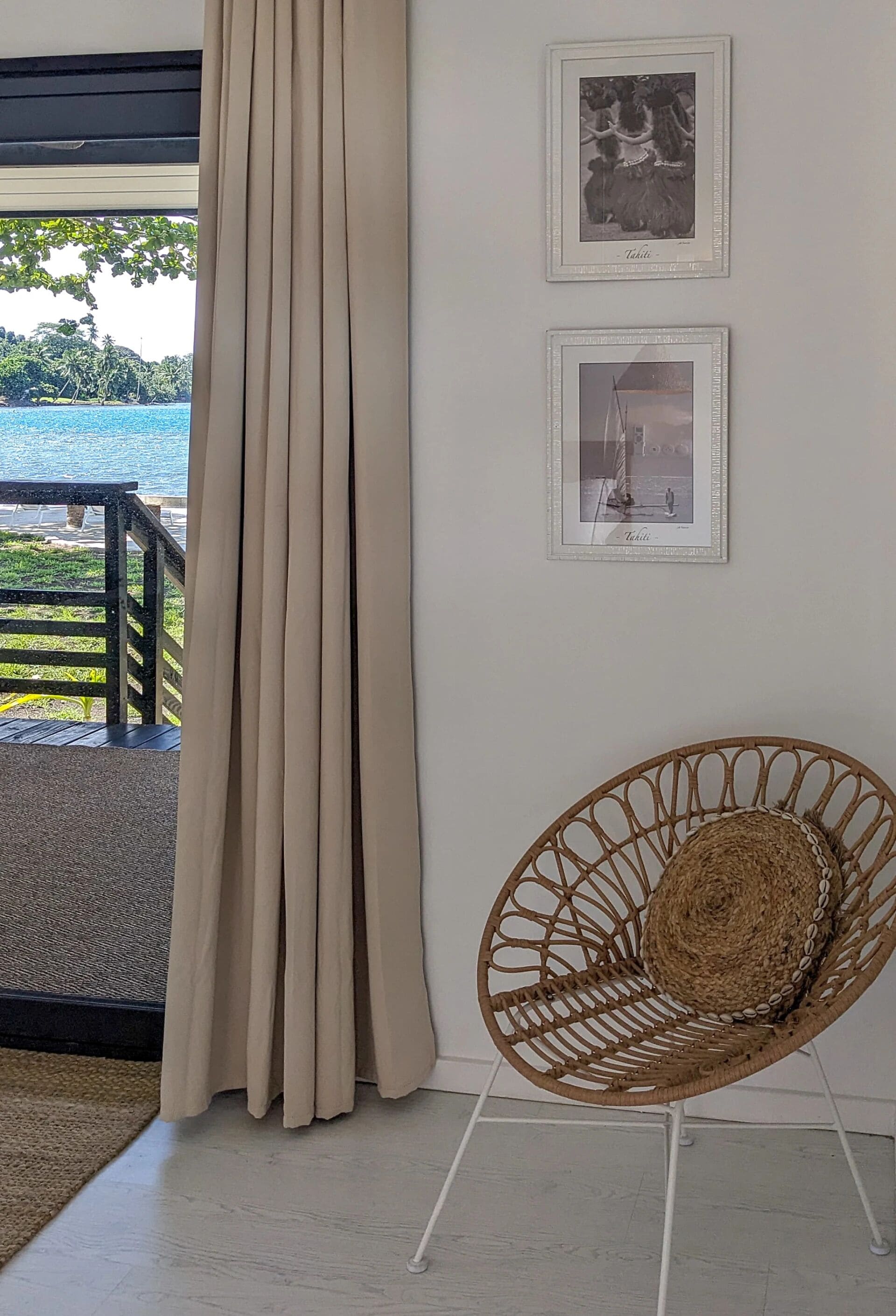 A room with a view of the sea, a wicker chair, and two framed pictures on the wall.