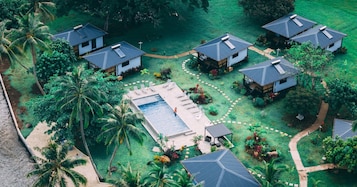 Resort com várias construções em estilo bangalô, piscina central e paisagem tropical exuberante.