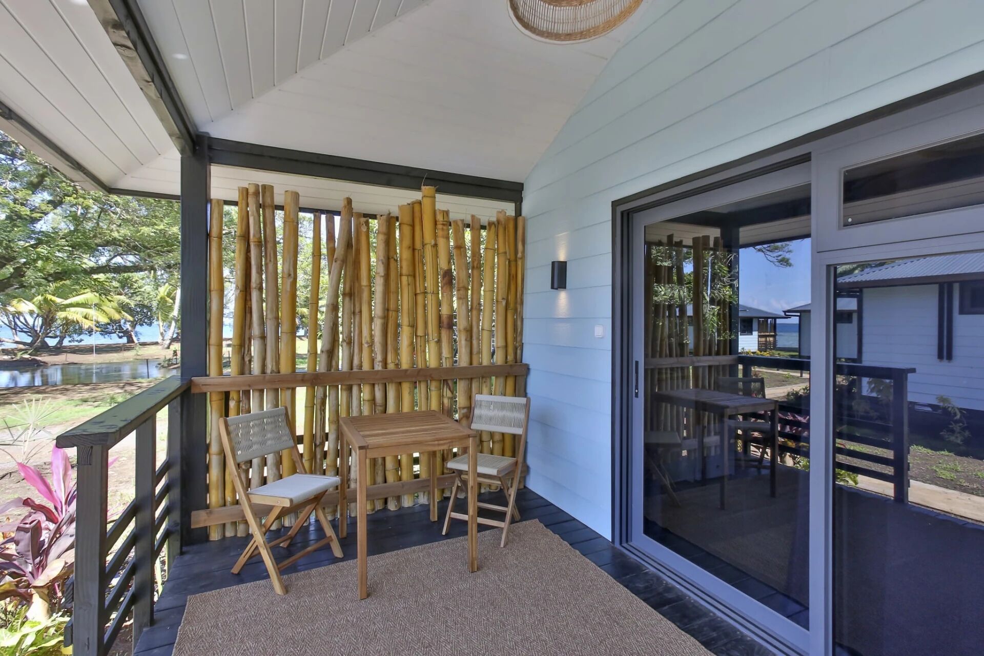 A covered outdoor area with a bamboo fence, a wooden table, two chairs, and a glass door leading to a pool area.