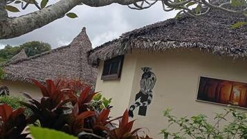 Una casa tradicional con techo de paja y un mural de una figura sentada, rodeada de abundante vegetación.