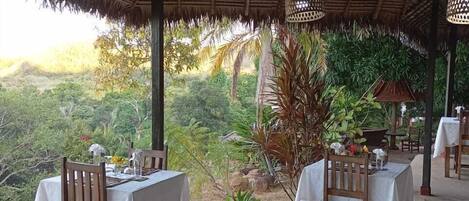 A covered outdoor dining area with tables and chairs, surrounded by lush greenery and a view of the landscape.