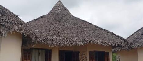 A bungalow with a thatched roof, a small porch, and two lounge chairs.