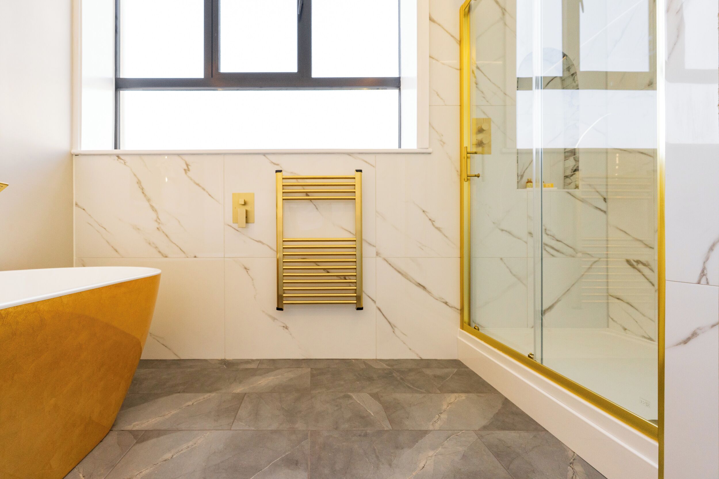 A modern bathroom with a marble wall, a gold bathtub, a glass shower enclosure, and a built-in towel rack.