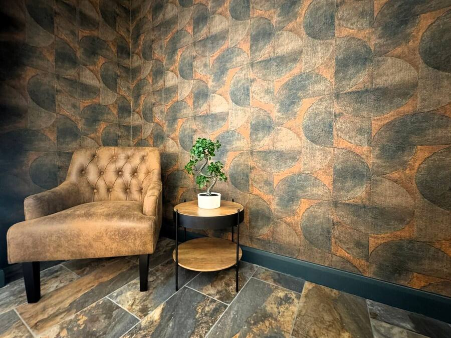 Un sillón con textura tufada y una mesa redonda de madera con una planta en maceta, frente a una pared con diseño y apariencia de piedra.