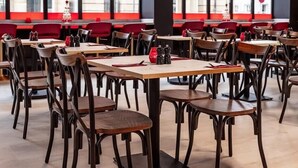 Um restaurante com várias mesas e cadeiras, janelas amplas com vista para edifícios e detalhes vermelhos nas paredes.