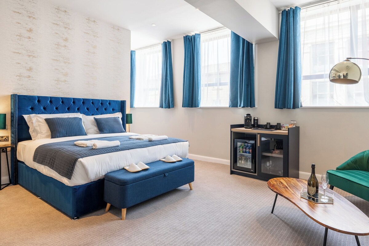 A modern hotel room with a large bed, a wooden coffee table, a minibar, and a green armchair.