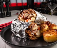 Comida que incluye un sándwich envuelto en papel de aluminio, con el relleno a la vista, y patatas asadas sobre un plato.