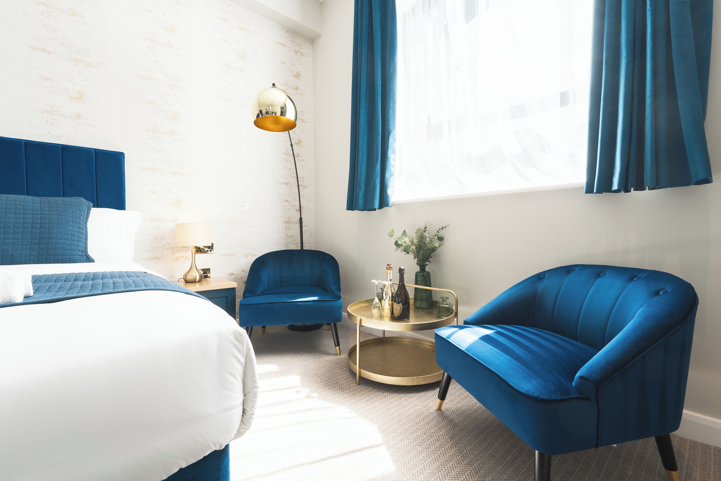 A hotel room with a bed, two blue armchairs, a small round table with a lamp, and a large window with curtains.
