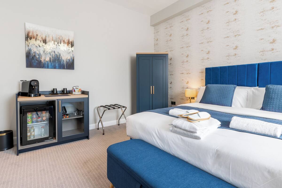 A hotel room with a bed, a minibar, a desk with a coffee machine, and a wall-mounted artwork.