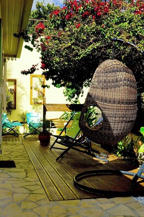 Une chaise en osier placée sous une pergola ornée de fleurs rouges, une terrasse en bois et des plantes en pot.