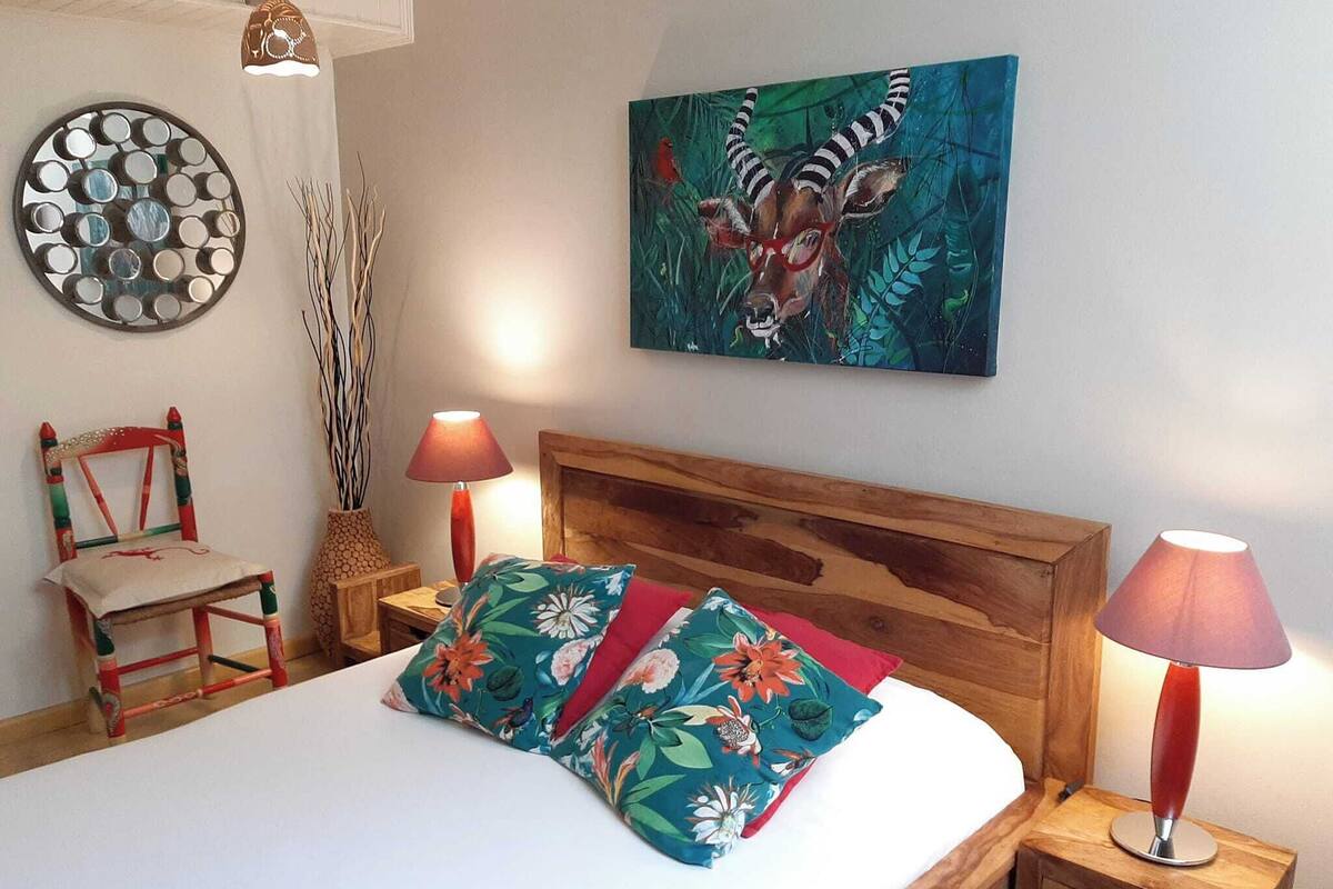 A bedroom with a wooden headboard, a bed with floral pillows, a wall art of a zebra, a decorative mirror, and a chair.