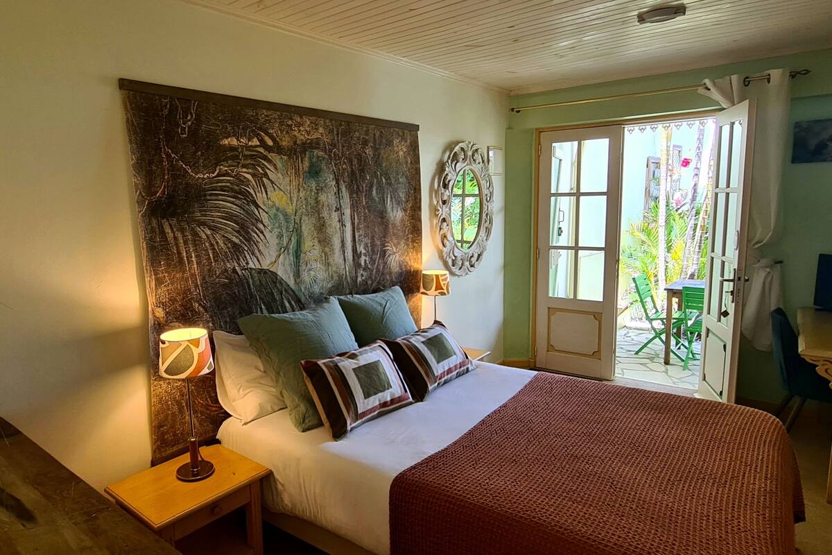 A bedroom with a large bed, two bedside tables with lamps, a decorative wall piece, and a view of the outside through an open door.