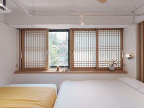 A bedroom with two beds, a window with wooden shoji screens, a bedside table with a vase, and a wall-mounted light fixture. - The Road Minihotel (Seoul)