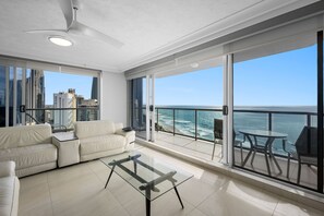 A modern living room with a glass table, a sofa, and a balcony with a view of the ocean and city skyline.