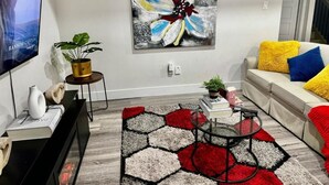 A modern living room with a large abstract painting, a sofa with colorful cushions, a coffee table, and a TV mounted on the wall. - Gateway to Serenity-welcome Home (London)