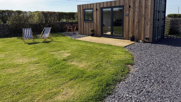 Una cabaña de madera moderna con puerta de vidrio, una pequeña terraza y dos sillones a rayas en un patio cubierto de césped.