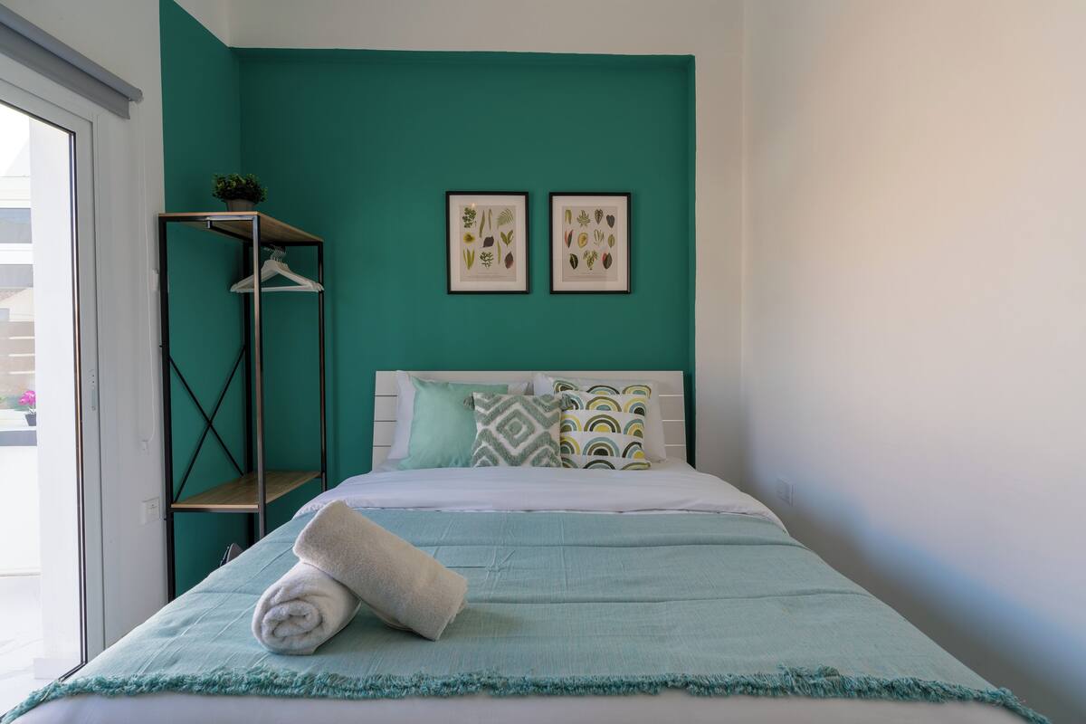 Un dormitorio ordenado con una pared azul verdoso, una cama con almohadones estampados, un estante colgante con una planta e ilustraciones botánicas enmarcadas.