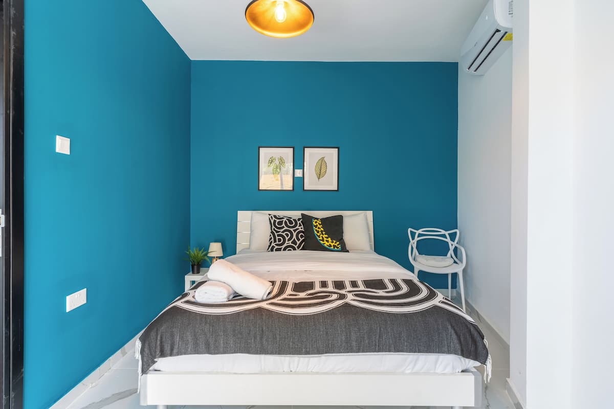 Un dormitorio con una pared color turquesa, una cama con colcha estampada, dos cuadros enmarcados, una silla blanca y un equipo de aire acondicionado.