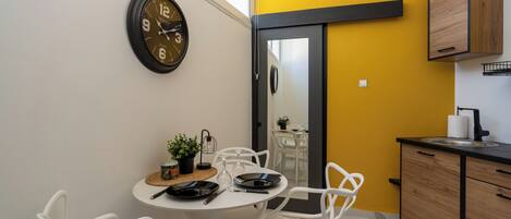 A compact kitchen with a dining area, featuring a clock, white chairs, a small table with a plant, and a yellow accent wall.