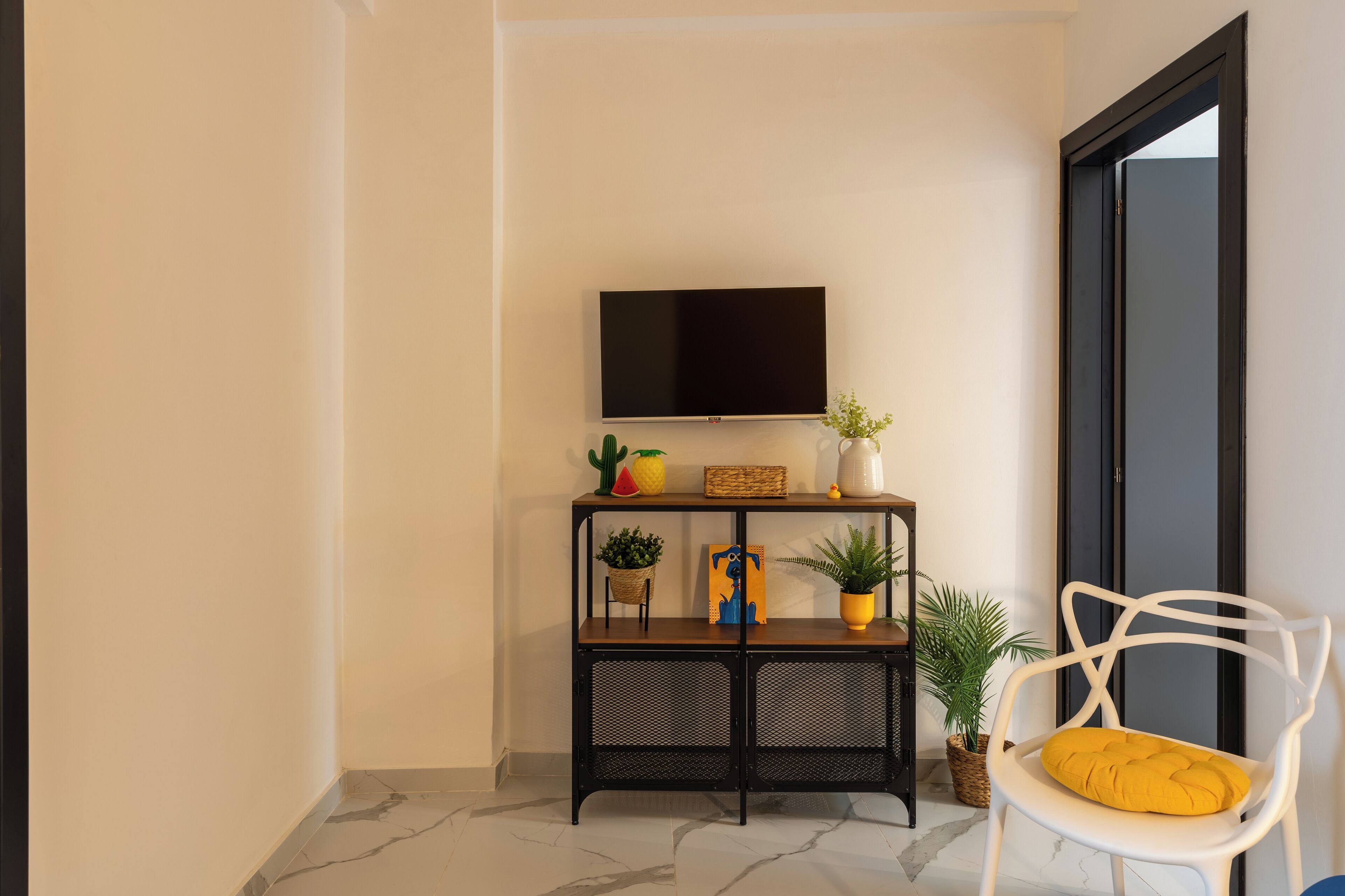 A modern living room with a flat-screen TV mounted on the wall, a black console table with decorative items, a white chair with a yellow cushion, and a large window with a view of the outside.