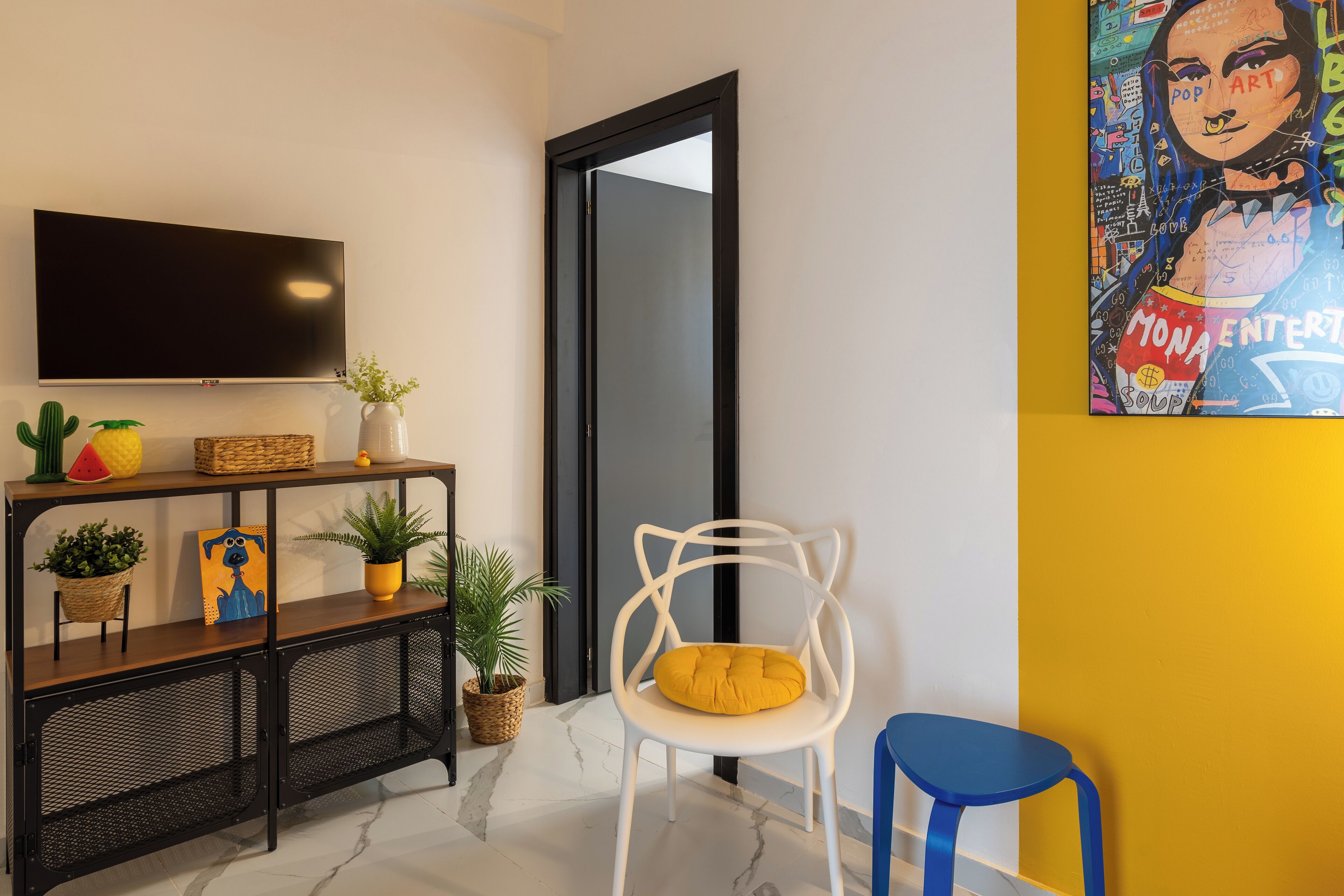 A modern living room with a flat-screen TV, a white chair with a yellow cushion, a blue stool, and a colorful painting on the wall.