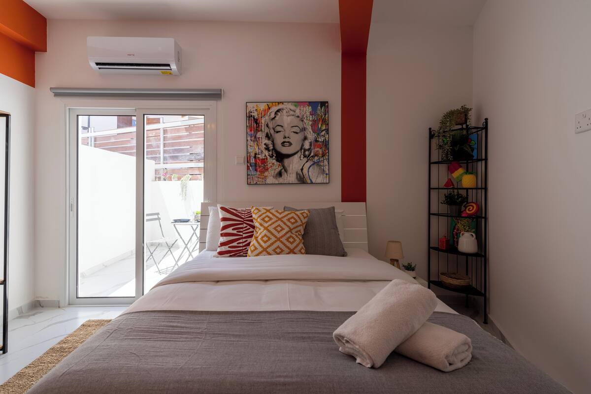 Un dormitorio moderno con una cama grande, un ventanal corredero a un balcón, una obra de arte colorida en la pared y un pequeño estante con objetos decorativos.