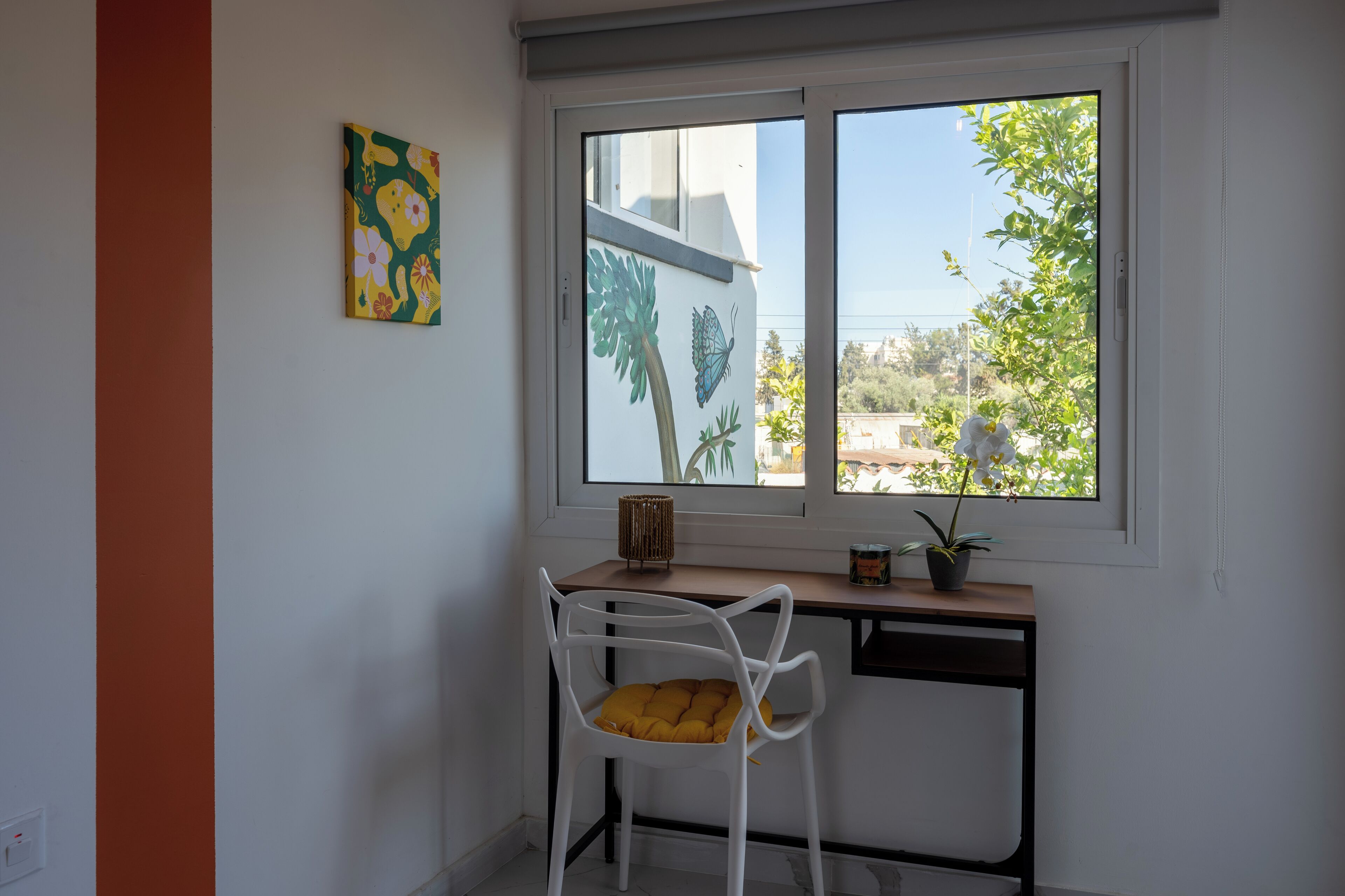 Una stanza con una scrivania, una sedia bianca con cuscino giallo, una finestra vista su del verde e un muro con un quadro colorato.