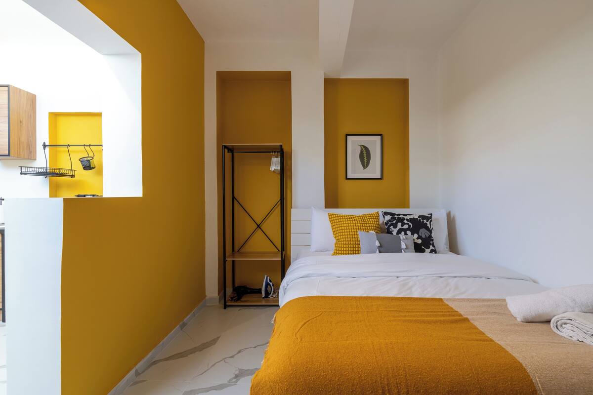 Una habitación de hotel con cama, pared amarilla, techo blanco y un cuadro en la pared.