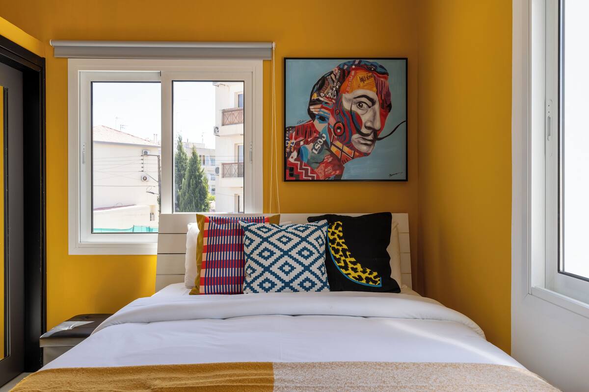 Habitación de hotel con cama, almohadas coloridas, un cuadro en la pared y ventana con vista a edificios.