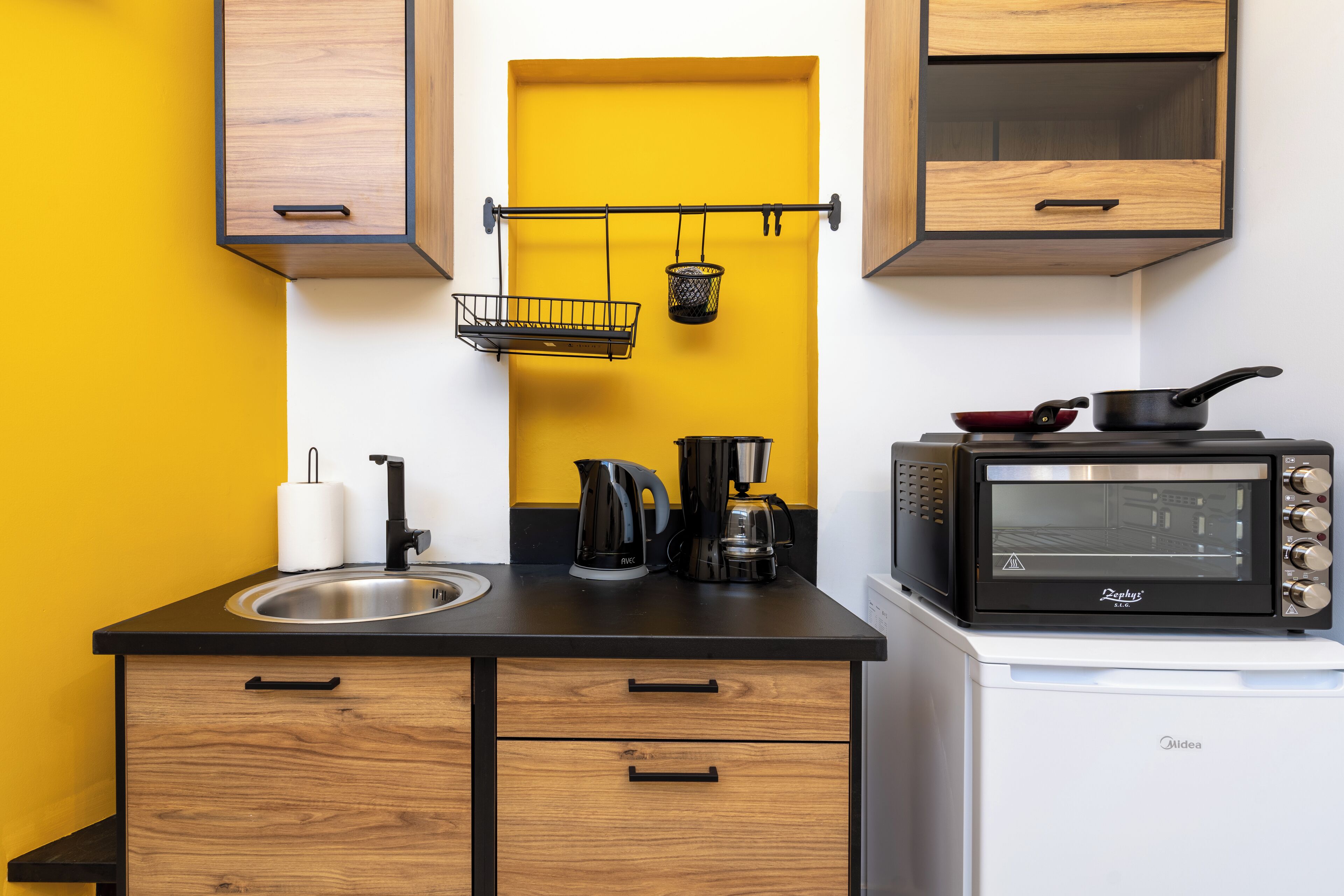 Eine kleine Küche mit schwarzem Arbeitsplatz, Holzschränken, einer gelben Akzentwand, einer Kaffeemaschine, einem Wasserkocher, einem Toaster und einem hängenden Korb.