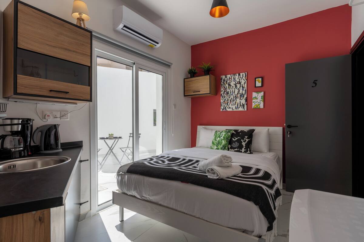 Un dormitorio moderno con una pared roja de acento, una cama grande, un armario empotrado, una zona de cocina con lavabo y cafetera, y un balcón con mesa y sillas.
