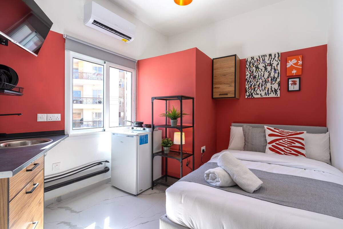 Habitación de hotel compacta con kitchenette, cama con ropa de cama blanca, pared de acento roja y ventana con vista a edificios.