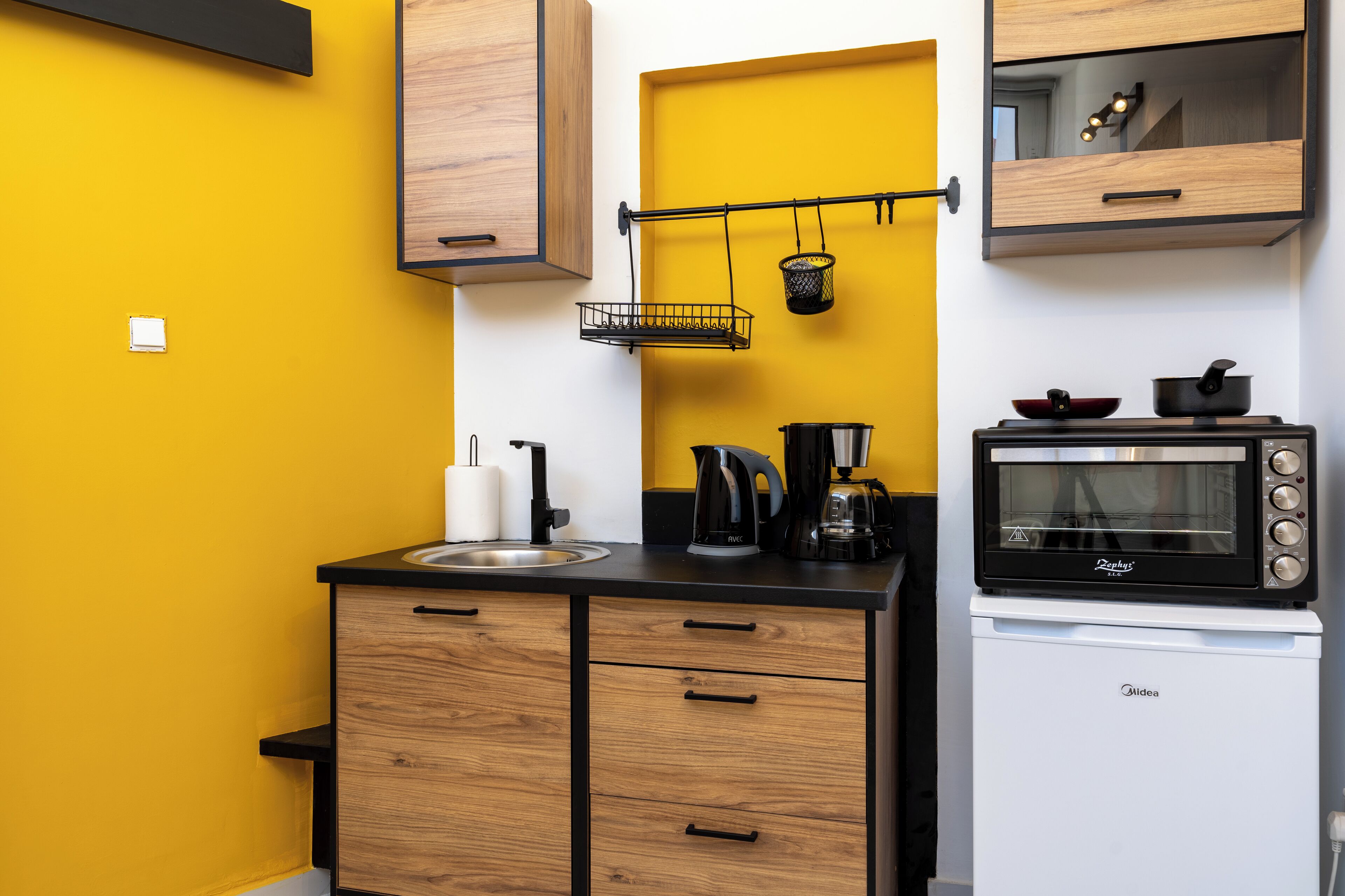 A compact kitchen with wooden cabinets, a black countertop, a coffee maker, a kettle, a microwave, and a hanging basket.