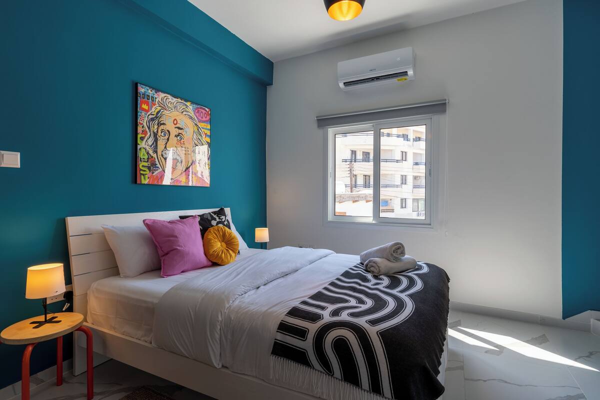 Un dormitorio con cama, mesita de noche, lámpara, ventana y aire acondicionado instalado en la pared.